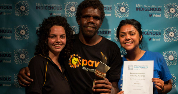 Three Indigenous broadcasters against background printed with 18th National Remote Indigenous Media Festival. Holding a certificate and an award.