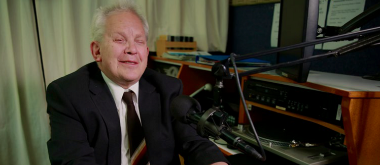 Vision impaired man in studio in front of microphone, operating a community radio console.