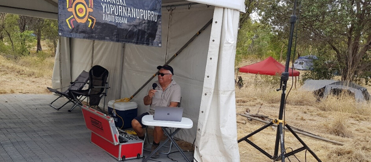 Seated older man, Wayne Bynder, with a cap and microphone broadcasting in a tent in the bush south east of Fitzroy Crossing