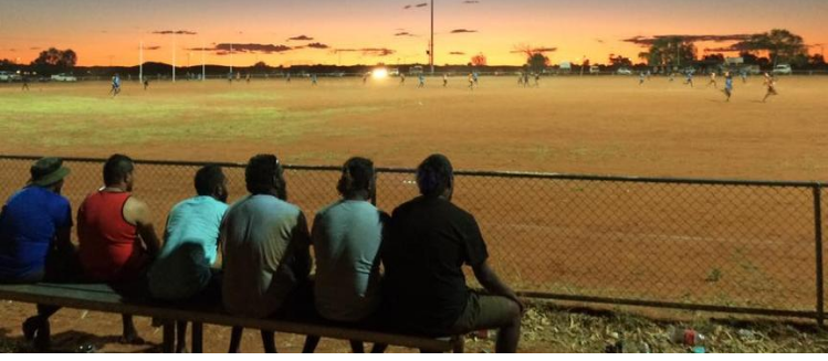 A row of men sitting on bench at sunset watching a football game in Yuendumu