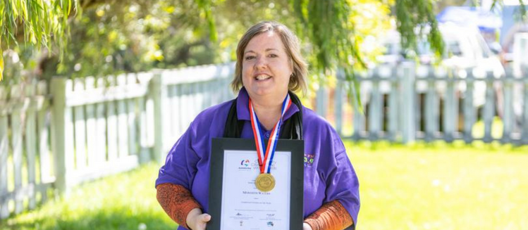 Woman in purple shirt holding framed certificate. Has a gold medal around her neck