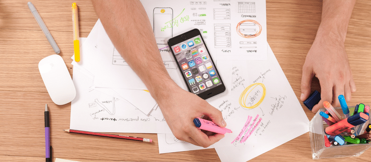 Arm reaching over desk of papers highlighting items. Mobile phone sitting on top of papers