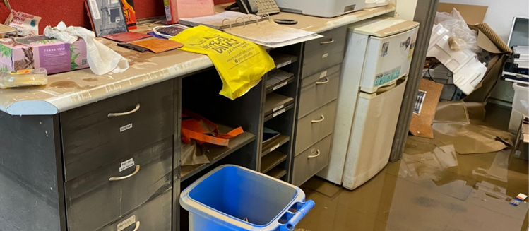 Room impacted by flood waters - papers and desk covered in dirty water. Blue bucket near by indicating a clean up