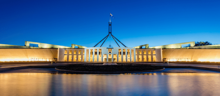 Parliament House in Canberra at dusk. White building has lights on glowing through the windows with an Australian flag flying above the building