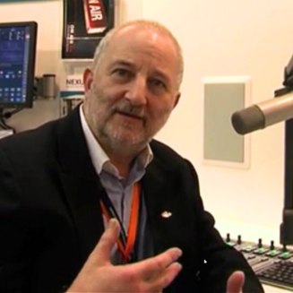 Man in a radio studio wearing a black suit jacket with light hair and gesturing mid explanation and leaning into studio mic