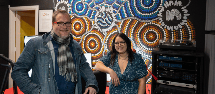 male with denim jacket and scarf and glasses smiling next to woman with glasses in navy patterned dress and both standing in front of wall featuring First Nations artwork in a studio