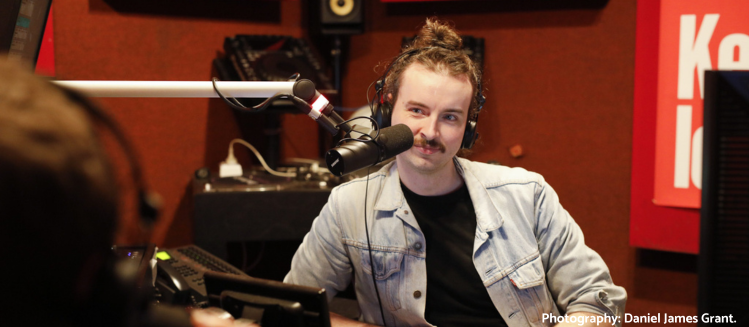 Male with light complexion and hair tied up in a bun with headphones on in the station studio