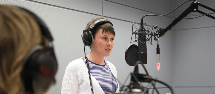 Person with short brown hair wearing a white cardigan and stripe ringer top with headphones one standing in front of studio microphone set up