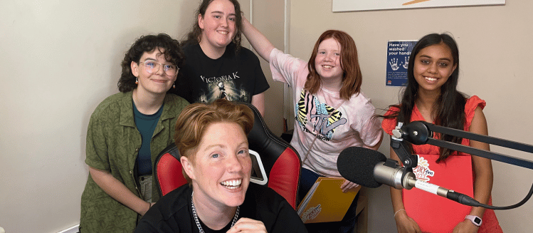 A group of young girls in khaki, black, pink and orange tops smiling in a radio studio and standing behind a woman adult with short red hair and black tee shirt and necklace