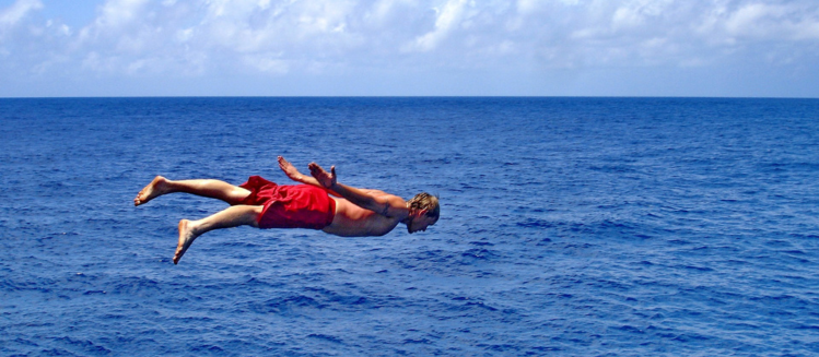 young man in bright red board shorts planking in the air and launching into the ocean before a cloudy bright blue sky