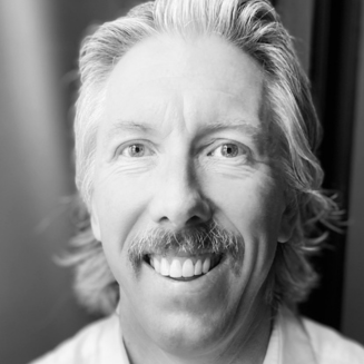 Close up black and white portrait of a man with moustache smiling at the camera in a collared shirt