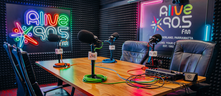 A sound proofed room featuring colour neon signs that read alive 90 point 5 fm and a wooden table with studio microphones and a portable mixer and leather chairs