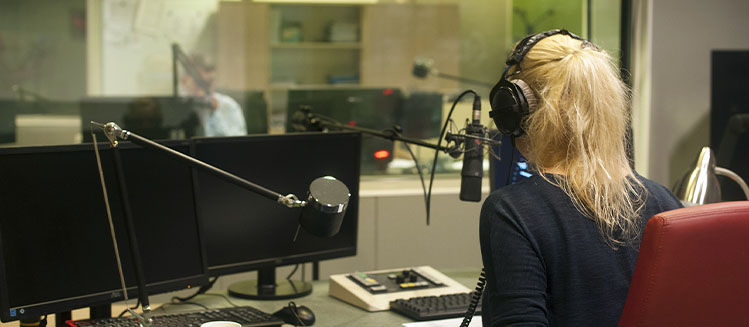 Back of a Blonde woman sitting in a studio in front of a microphone and mixing console with headphones on