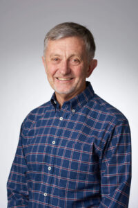 Man smiling at camera with grey hair and grey stubble wearing blue checked shirt