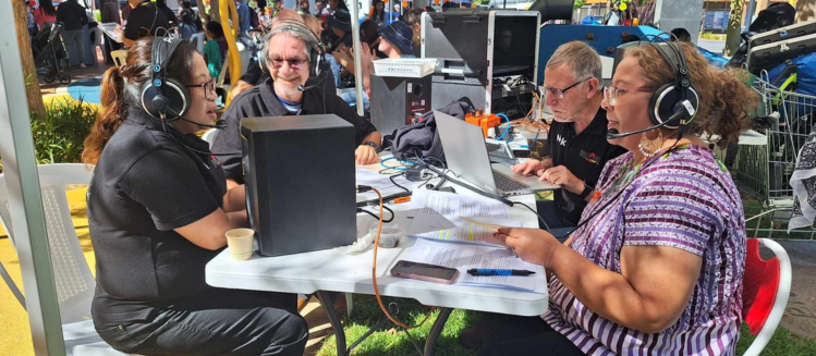 A group of four people of diverse gender and ethnicities sitting at a table wearing headphones conducting a live outdoor broadcast for an ethnic programming station for Harmony Day