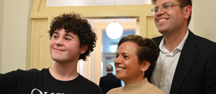 Young boy with dark hair smiling next to woman with short hair and beige top and a man wearing glasses in a black suit with white collared shirt