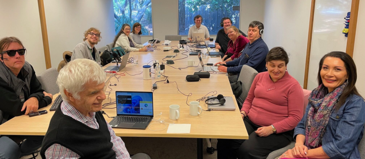 A group of people with vision impairment sitting at a long wooden table in a training conference room with laptops and microphones