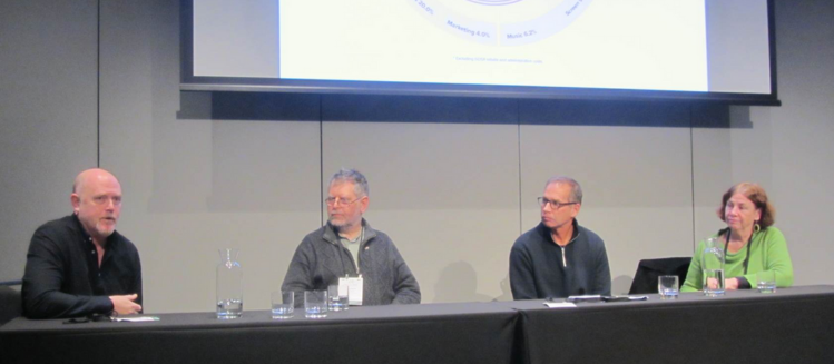 Three men wearing black grey and navy long sleeve tops and a woman wearing bright green sitting on a panel below a screen