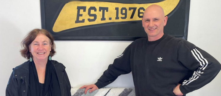 A woman smiling in a black coat next to man smiling in a striped sports jumper posing in front of a white wall with a yellow sign that reads est 1976