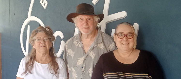 A woman with light coloured long hair wearing a white tshirt next to a man with a brown hat and collared shirt standing next to another woman wearing a black top with glasses standing outdoors in front of a dark wall