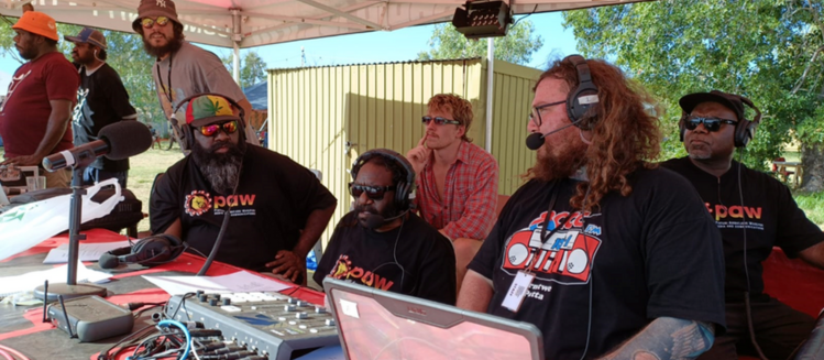 A group of eight people sitting at an outside broadcast gathered around a desk with a mixing console and broadcast equipment