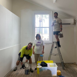 three people standing inside a white room with painting equipment and gear with one person standing on a step ladder