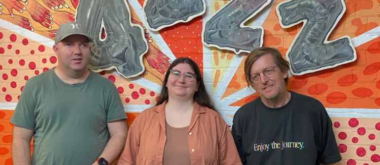 Two men and a woman standing outdoors in front of an orange wall with the words 4ZZZ