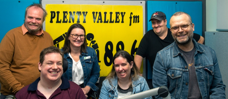 A group of people smiling and gathered in a radio station studio in front of a bright yellow banner on the wall that reads PLENTY VALLEY FM